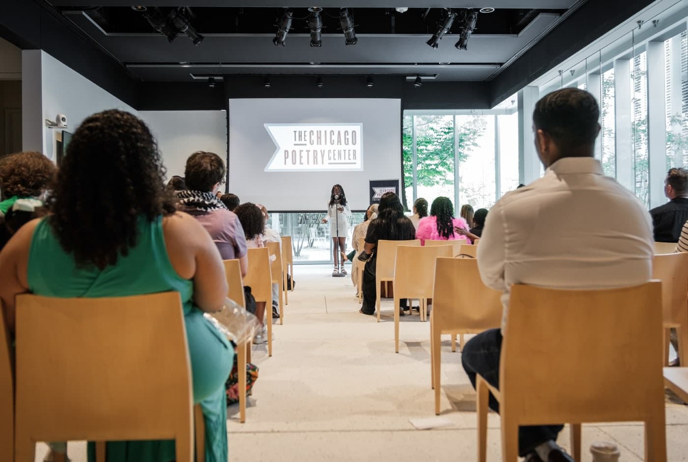 View from the back of an audience watching a young student perform at a microphone during an All Schools Reading, with the Chicago Poetry Center logo displayed on a screen and natural light streaming through floor-to-ceiling windows.