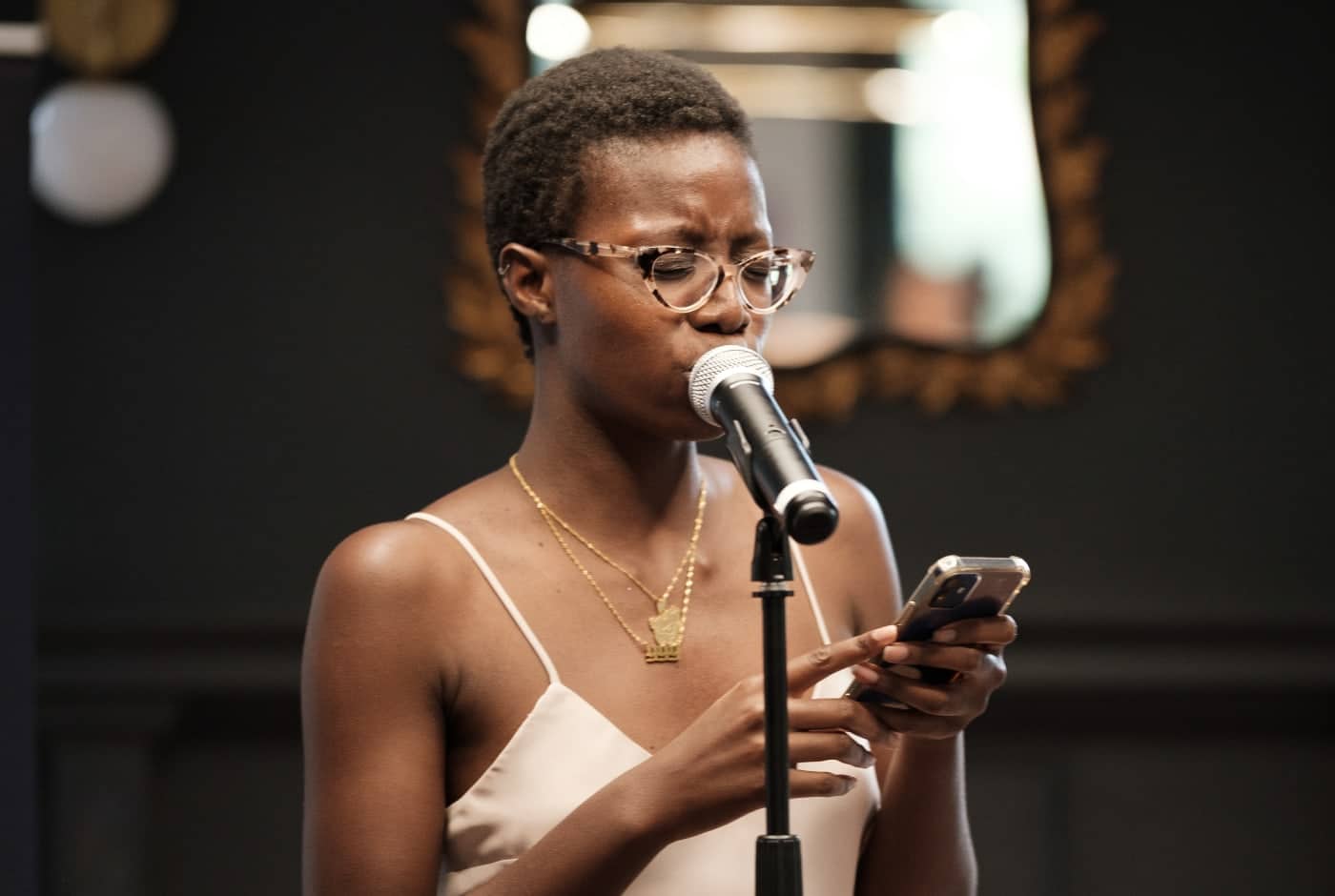 A poet in a cream-colored tank top and tortoiseshell glasses reads from her phone at a microphone during a Blue Hour reading, with warm ambient lighting in the background.