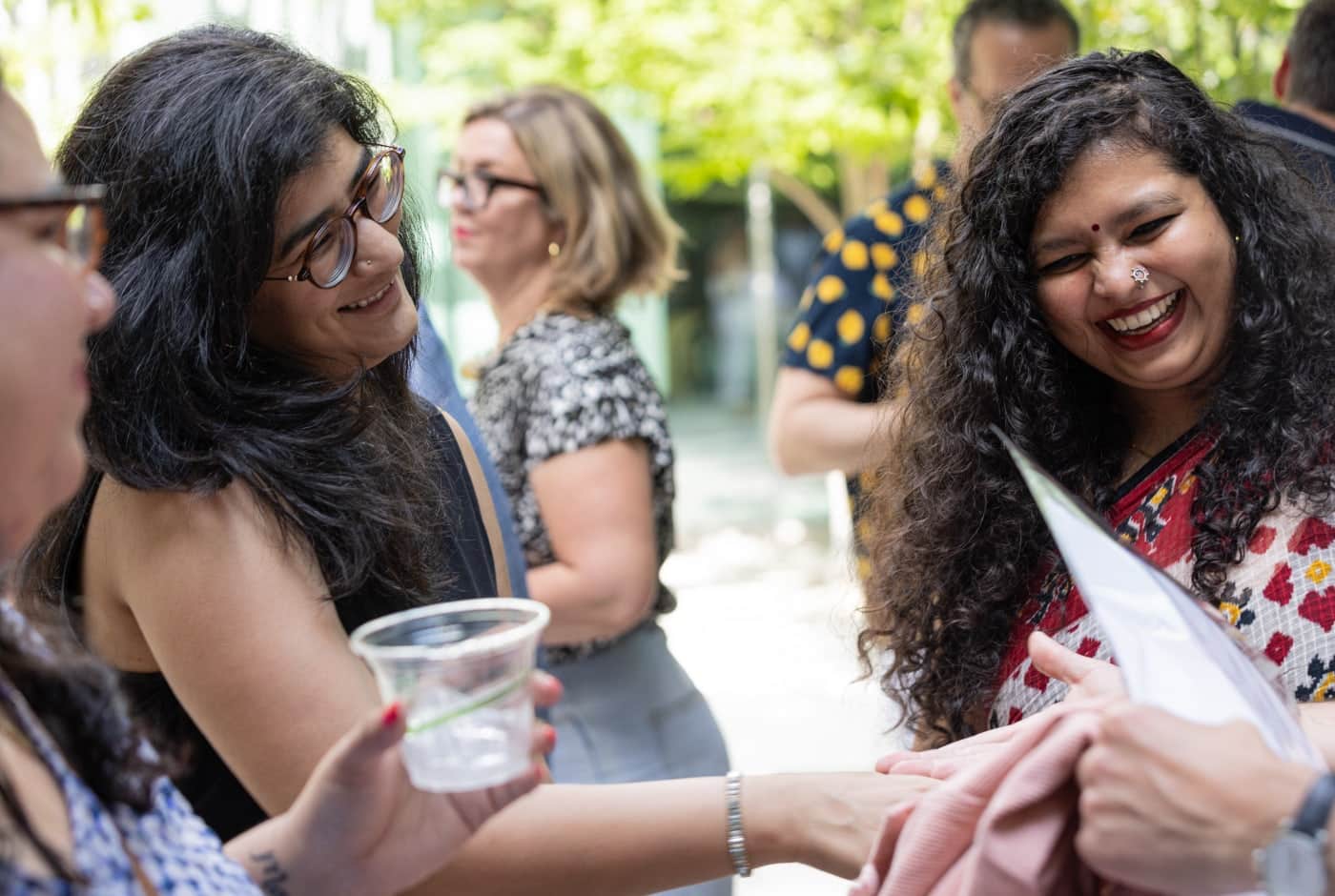 Two women share a laugh at an outdoor poetry event reception, one holding a drink, with other attendees mingling in the sunlit background.