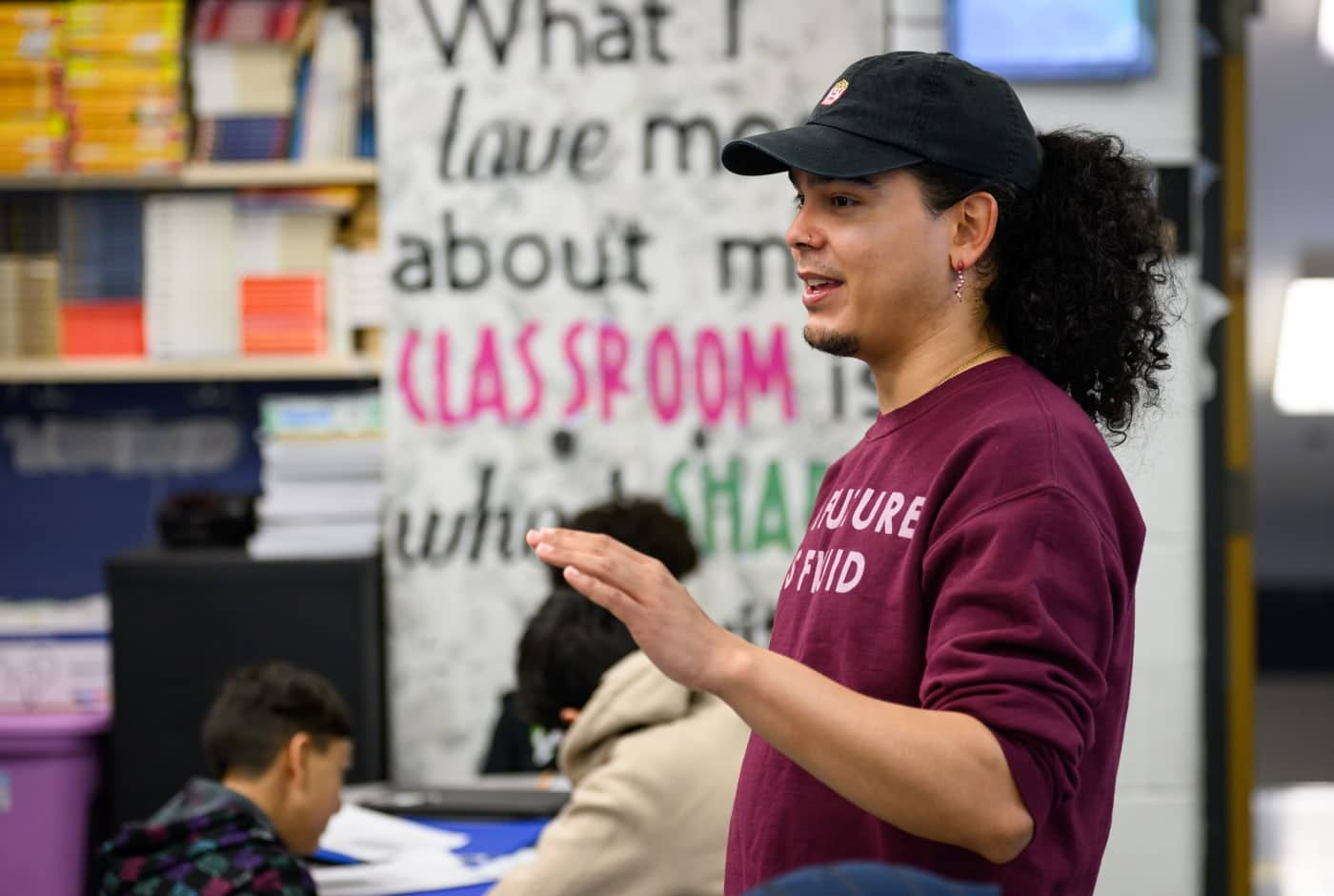 A teaching artist in a maroon sweatshirt and cap gestures while leading a lesson, with a colorful "What I love most about my classroom" poster visible behind them.