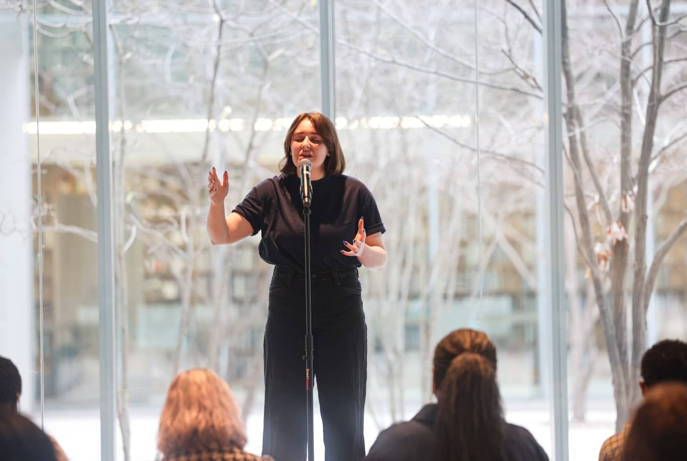 A young woman in all black performs poetry at a microphone, hands gesturing expressively, in front of floor-to-ceiling windows revealing bare winter trees. Audience members watch from the foreground.