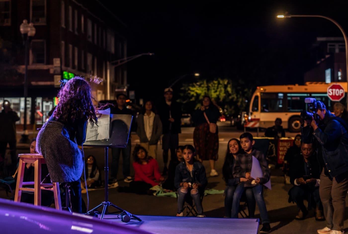 A poet reads from a music stand on a purple-lit outdoor stage at night in the Pilsen neighborhood, with children and adults gathered on the street to listen.