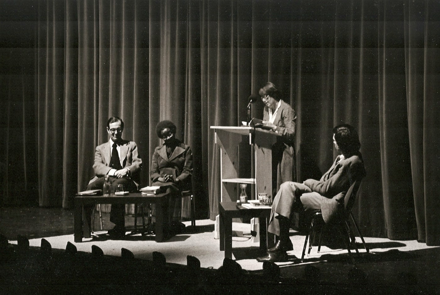 Black-and-white archival photo of a poetry reading on stage: a woman reads at a podium while three others sit nearby, with dramatic stage lighting against a curtained backdrop.