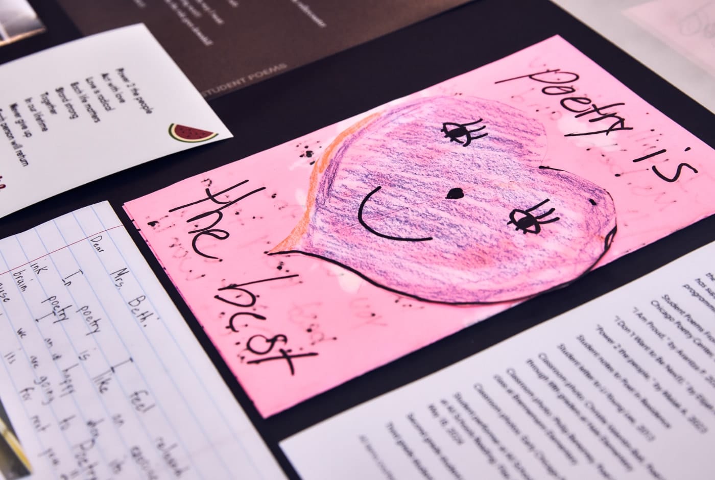 A child's handmade pink card featuring a crayon drawing of a smiling face and the words "Poetry is the best," displayed alongside handwritten letters and printed student poems.