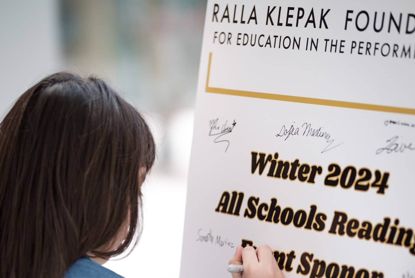 A student signs a sponsor recognition poster for the Winter 2024 All Schools Reading, presented by the Ralla Klepak Foundation, joining other handwritten signatures on the board.