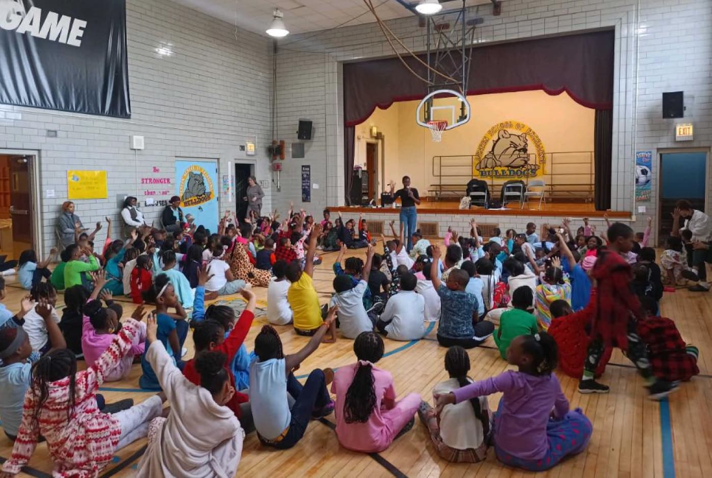 Dozens of elementary students sit on a gymnasium floor with hands raised during a poetry assembly, facing a stage with presenters and a "Bulldogs" school mascot banner.