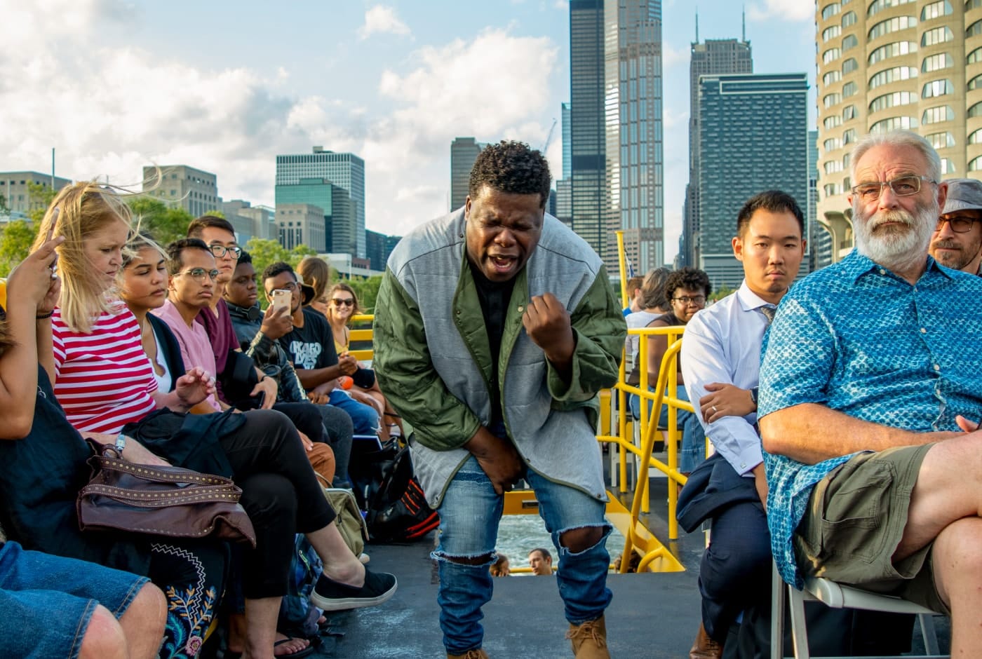 A poet delivers an impassioned performance on the deck of a boat, fists clenched and leaning forward with intensity, as audience members watch closely with the Chicago skyline rising behind them.