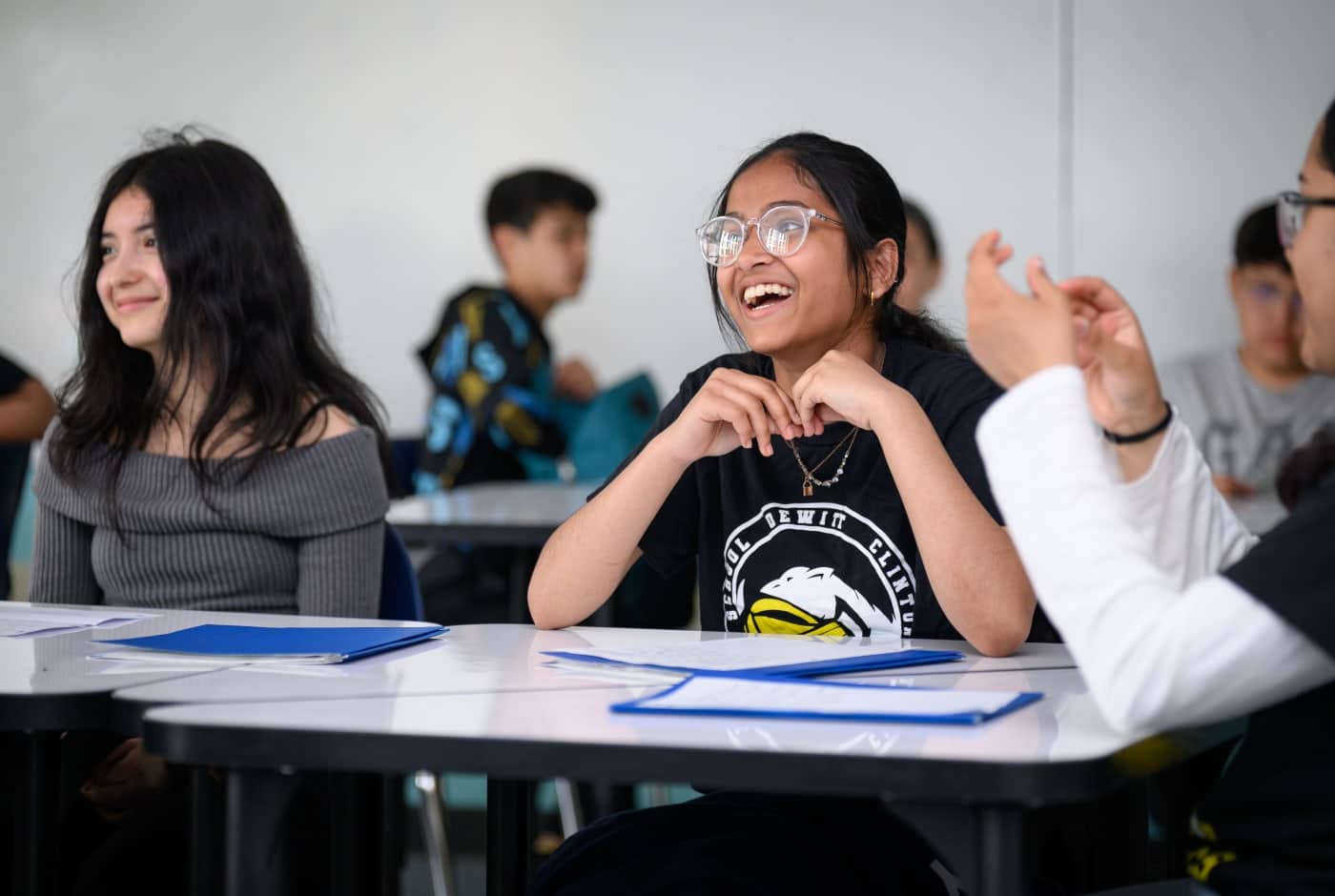 Middle school students laugh and engage during a poetry lesson, with blue folders on their desks and classmates working in the background.