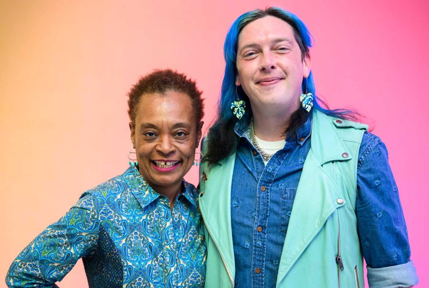 Two staff members smile together against a vibrant pink-to-orange gradient background. One wears a blue paisley shirt with turquoise earrings; the other has long blue hair and wears a mint top layered with a denim vest.