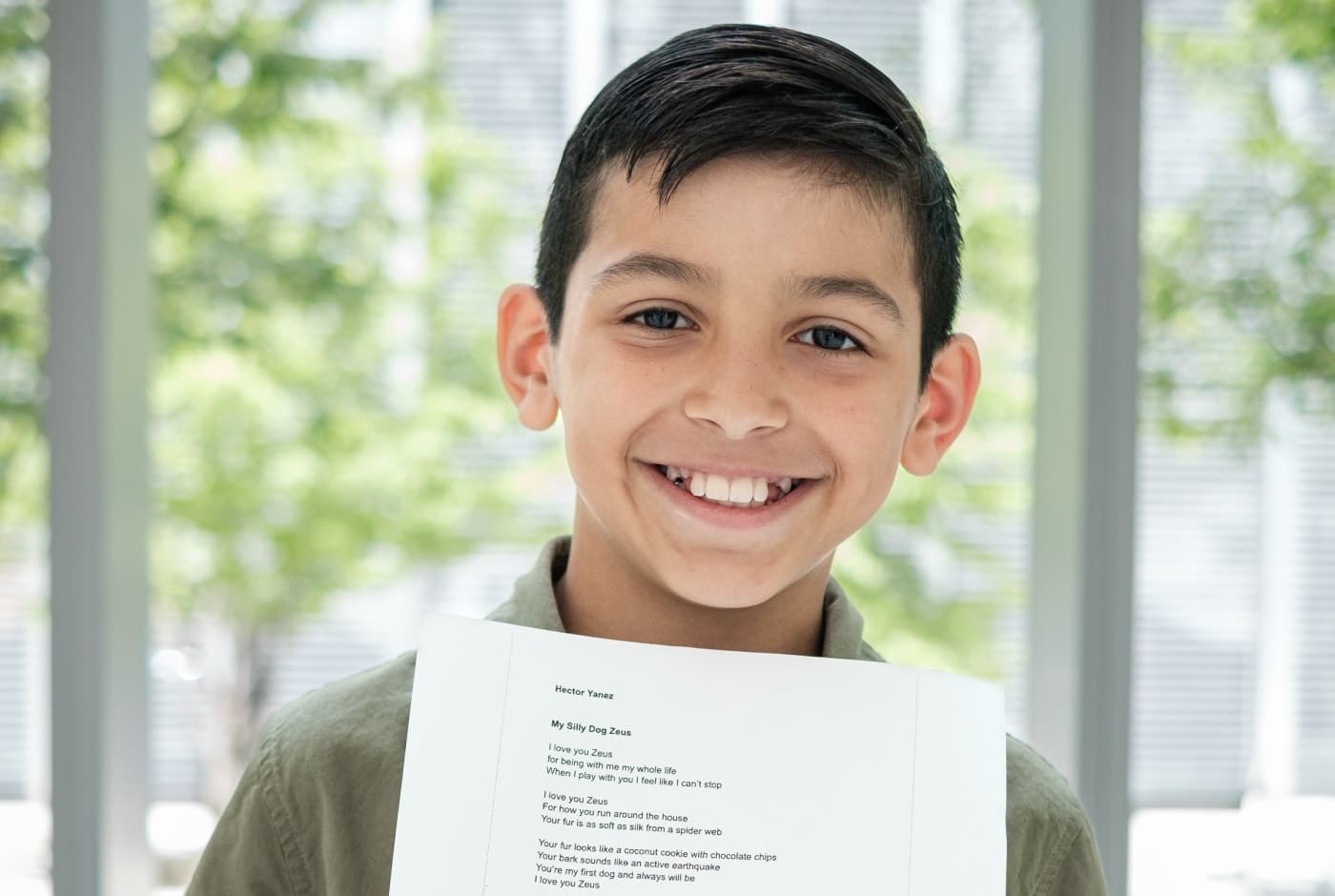 A young student beams with pride while holding up a printed copy of his poem titled "My Silly Dog Zeus," standing in front of a window with greenery visible outside.